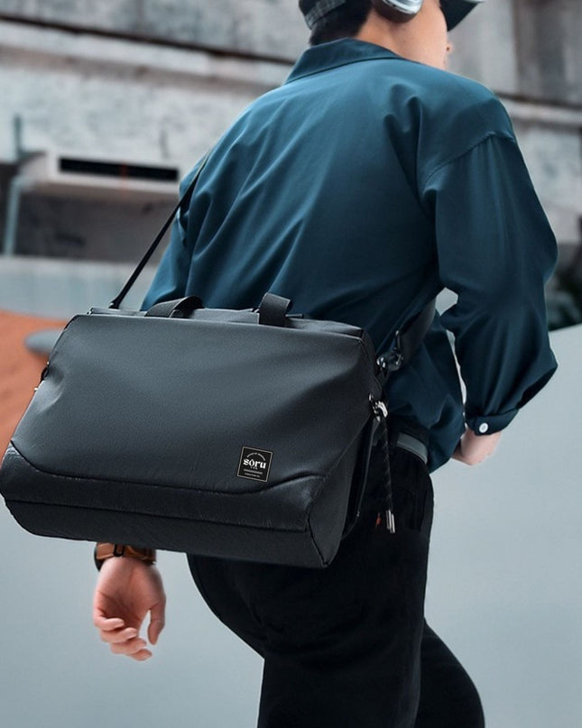 Person wearing a dark green jacket with a black bag over their shoulder, standing on a concrete surface.